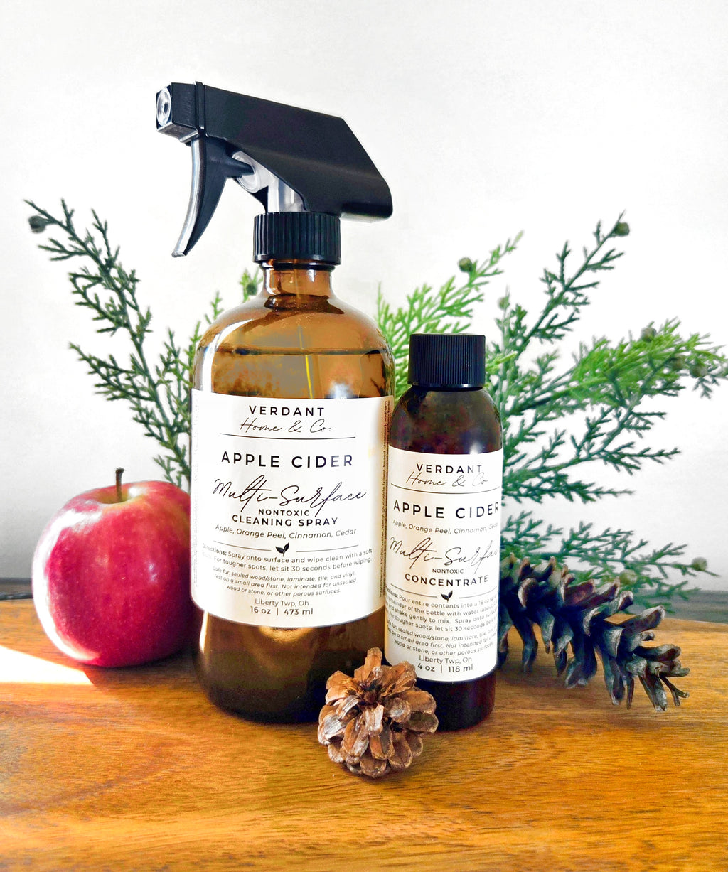 Two amber glass cleaning spray bottles from Verdant Home & Co. on a wooden surface, styled with a red apple, pinecones, and green pine branches against a white background.