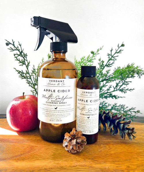 Two amber glass cleaning spray bottles from Verdant Home & Co. on a wooden surface, styled with a red apple, pinecones, and green pine branches against a white background.