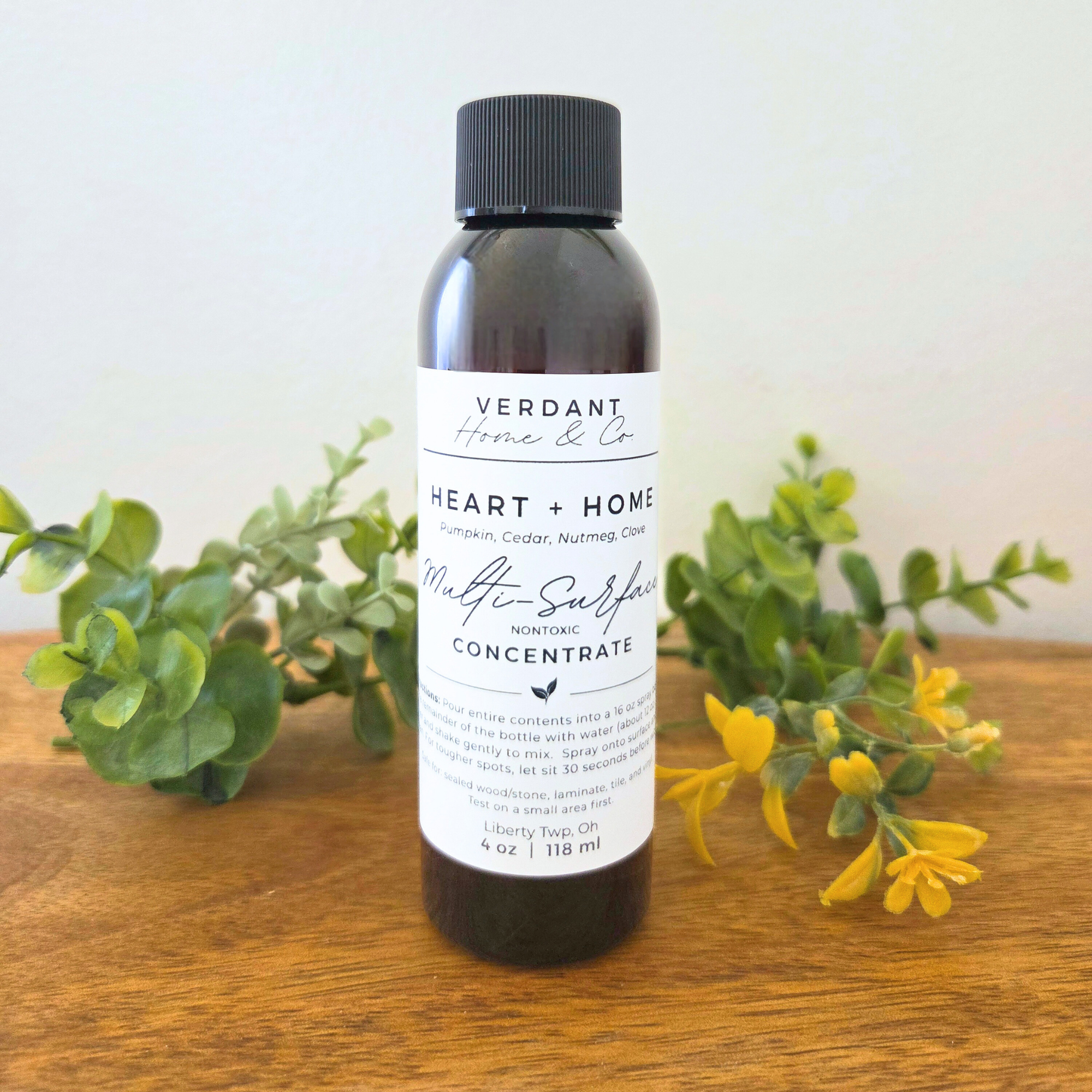 Bottle of Verdant Home & Co concentrate on a wooden surface with greenery and flowers.