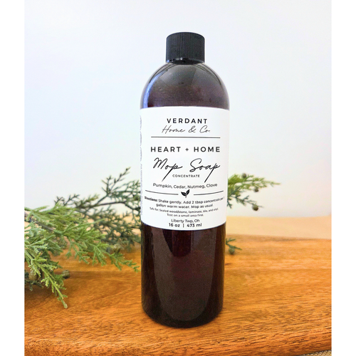 Bottle of Verdant Home & Co. Heart + Home Mop Soap Concentrate on a wooden surface with greenery in the background.