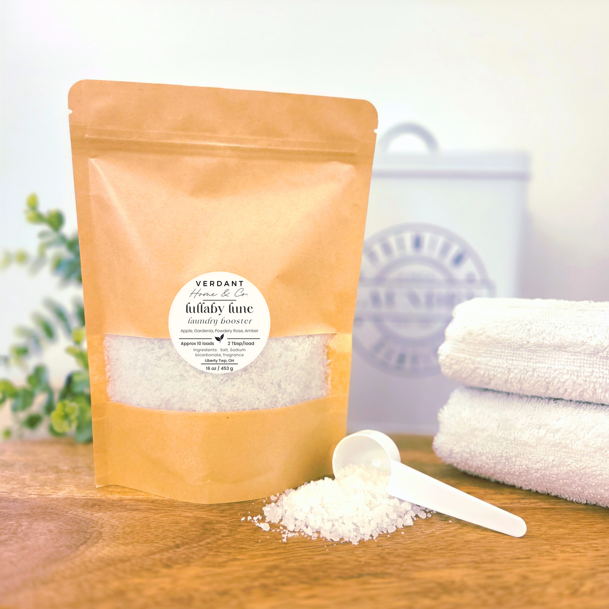 Bag of Verdant Home & Co laundry booster in scent Lullaby Lune with a scoop and towels on a wooden surface