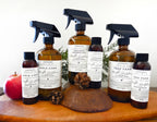 Six amber glass cleaning spray bottles with black spray tops and white labels from Verdant Home & Co, arranged on a wooden surface with a red apple and pinecones as props.
