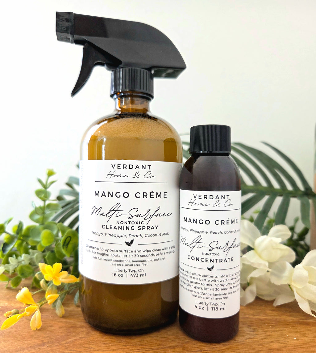 Two bottles of Verdant Home & Co Mango Crème Multi-Surface Cleaning Spray on a wooden surface with plants in the background.