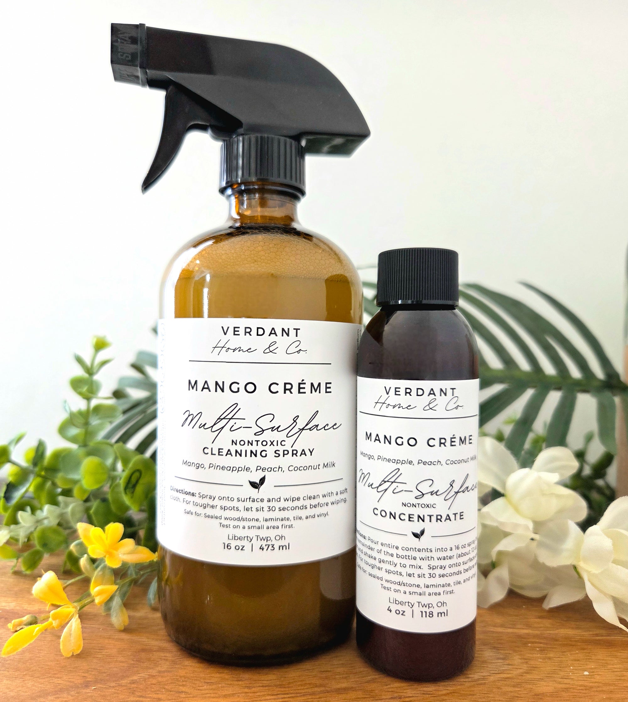 Two bottles of Verdant Home & Co Mango Crème Multi-Surface Cleaning Spray on a wooden surface with plants in the background.