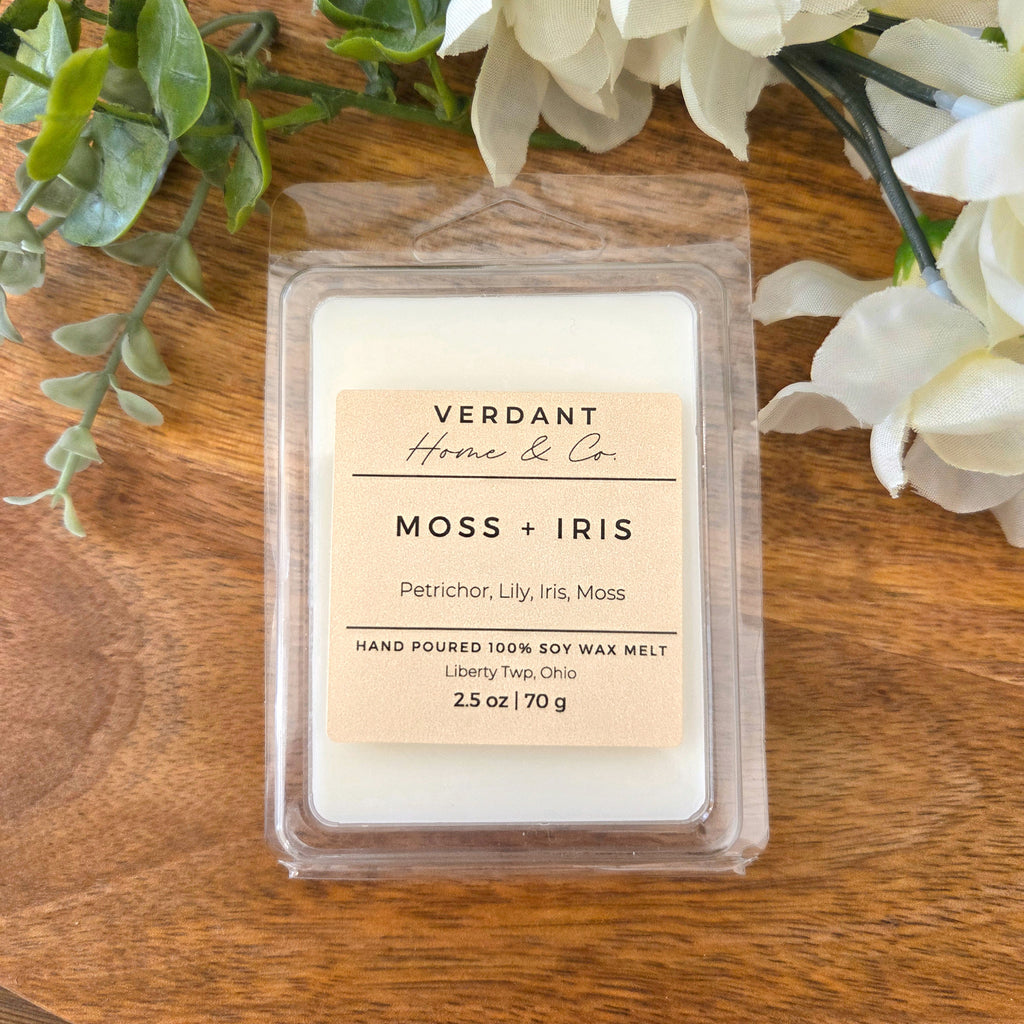 Verdant Home & Co. Moss + Iris wax melt on a wooden surface with flowers and greenery.