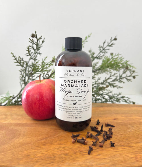 A bottle of Verdant Home & Co. Orchard Marmalade Mop Soap Concentrate stands on a wooden surface with a red apple and greenery in the background.