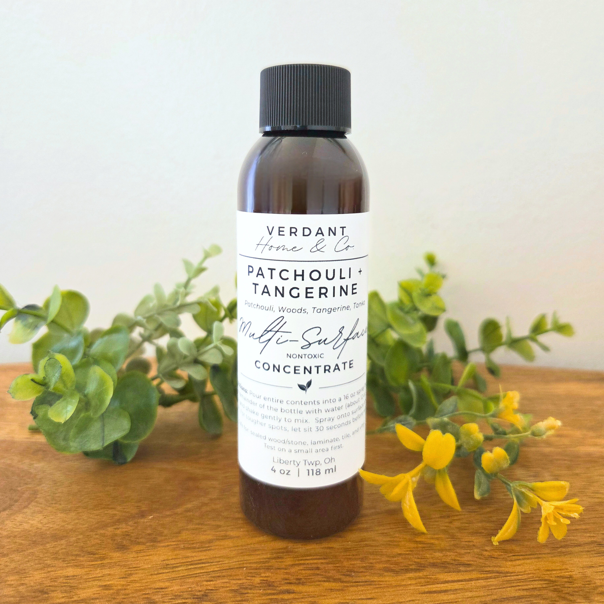 Bottle of Verdant Home & Co Patchouli + Tangerine product on a wooden surface with greenery and flowers.
