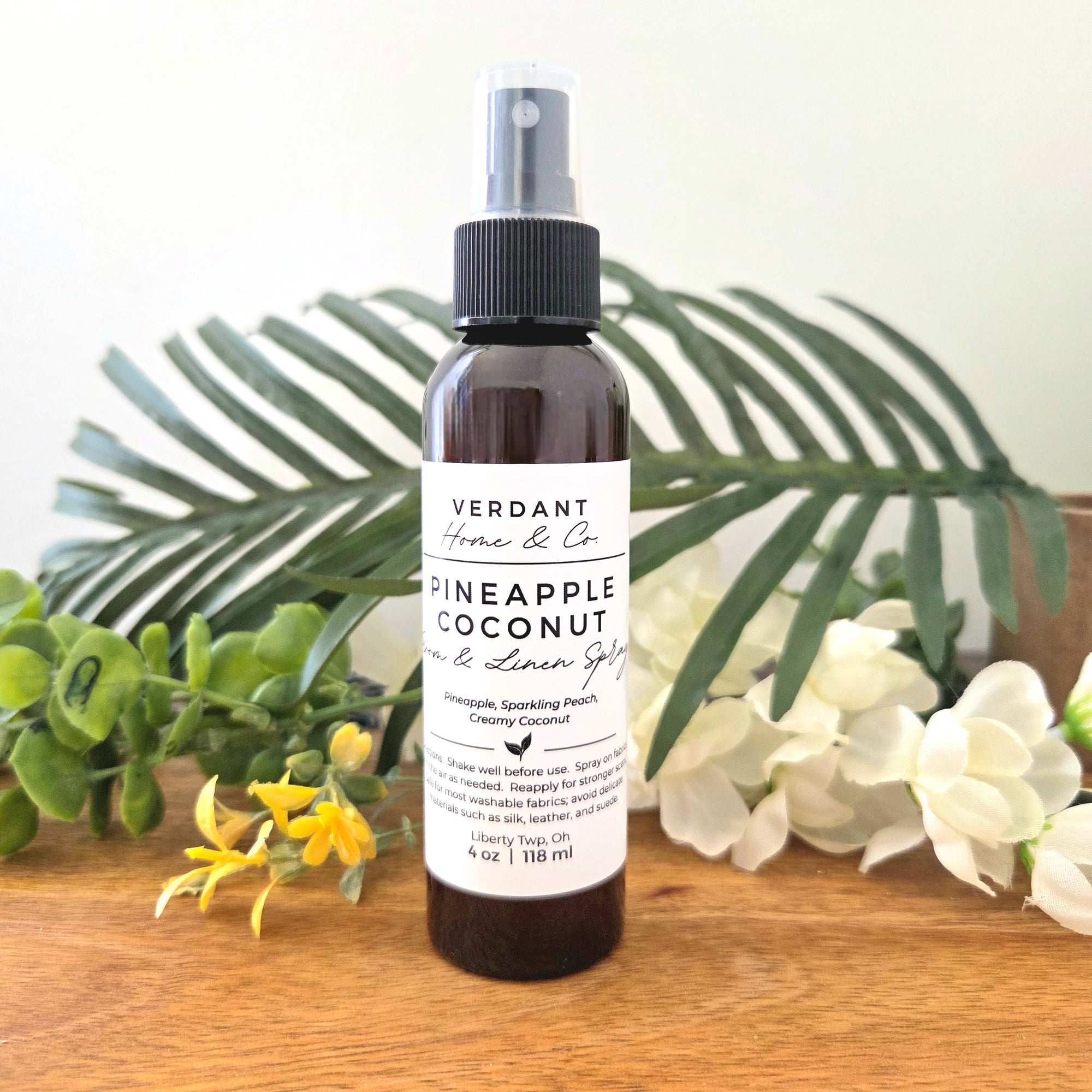 Bottle of Verdant Pineapple Coconut linen spray on a wooden surface with greenery and flowers in the background