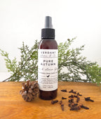 Brown bottle of Verdant Home & Co's room spray in the scent Pure Autumn, sitting on a wooden base with a pine cone and cloves sitting next to it.