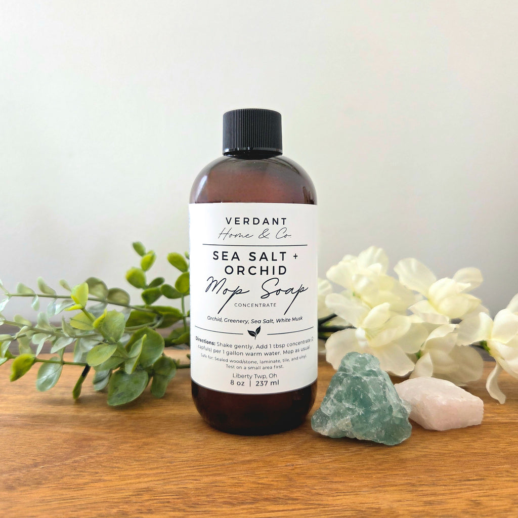 Bottle of Verdant Home & Co. Sea Salt + Orchid Mop Soap on a wooden surface with plants and stones.