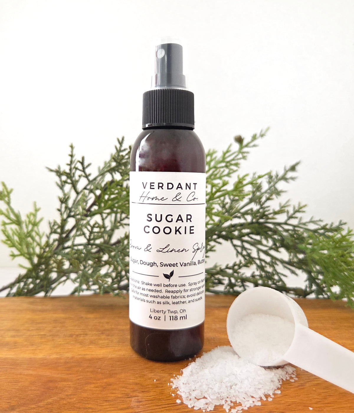 Brown bottle of Verdant Home & Co's room spray in the scent Sugar Cookie, sitting on a wooden base with sugar crystals next to it.