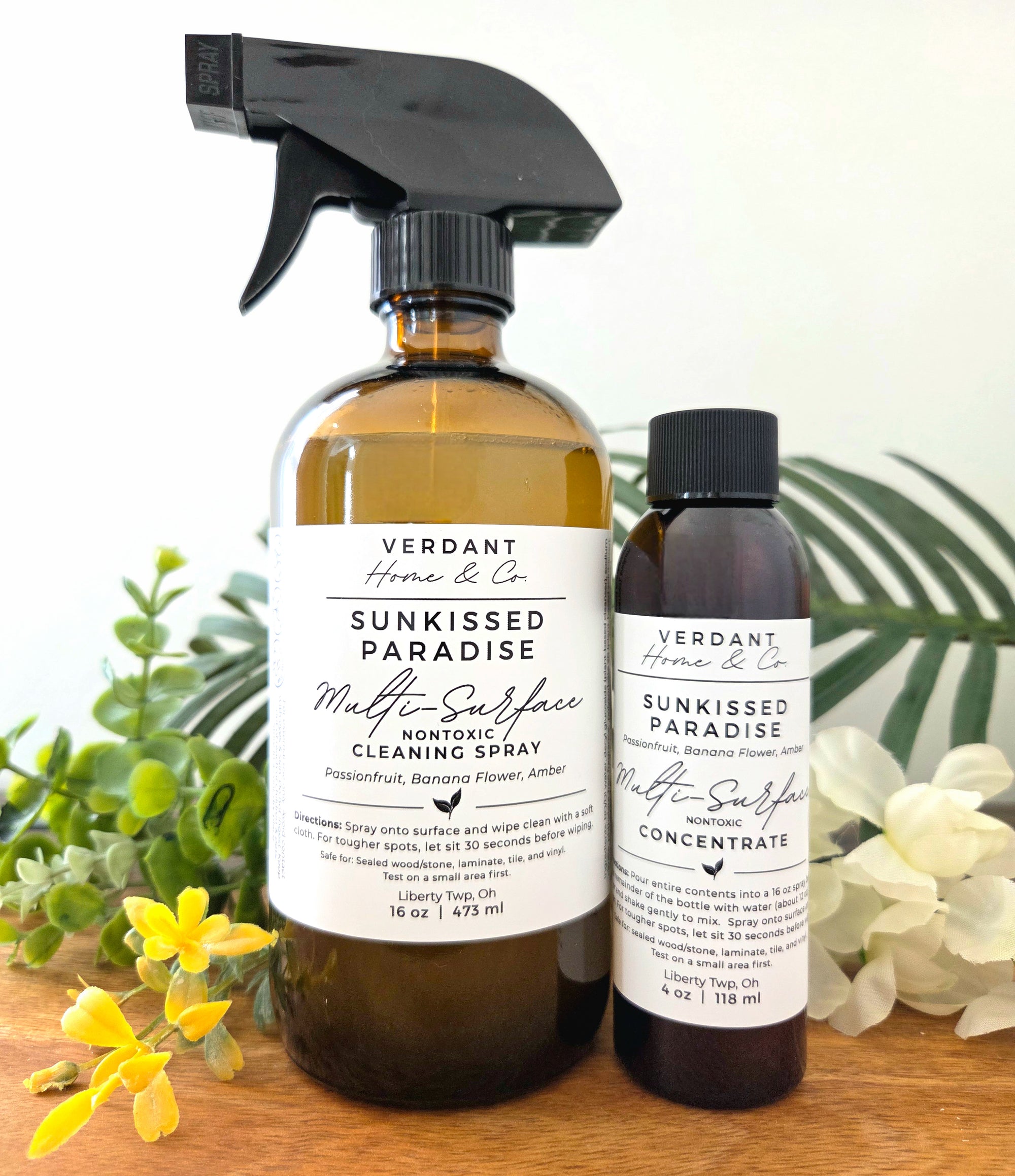 Two bottles of Verdant Home & Co. Sunkissed Paradise Multi-Surface Cleaning Spray and Concentrate on a wooden surface with plants in the background.