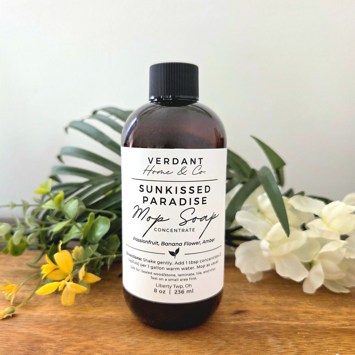 Bottle of Verdant Home & Co Sunkissed Paradise Mop Soap on a wooden surface with flowers and leaves.