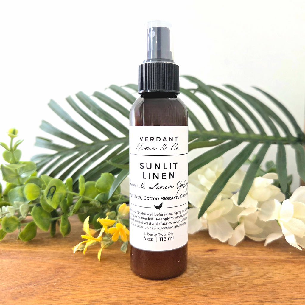 Bottle of Verdant Home & Co Sunlit Linen spray with greenery and flowers in the background