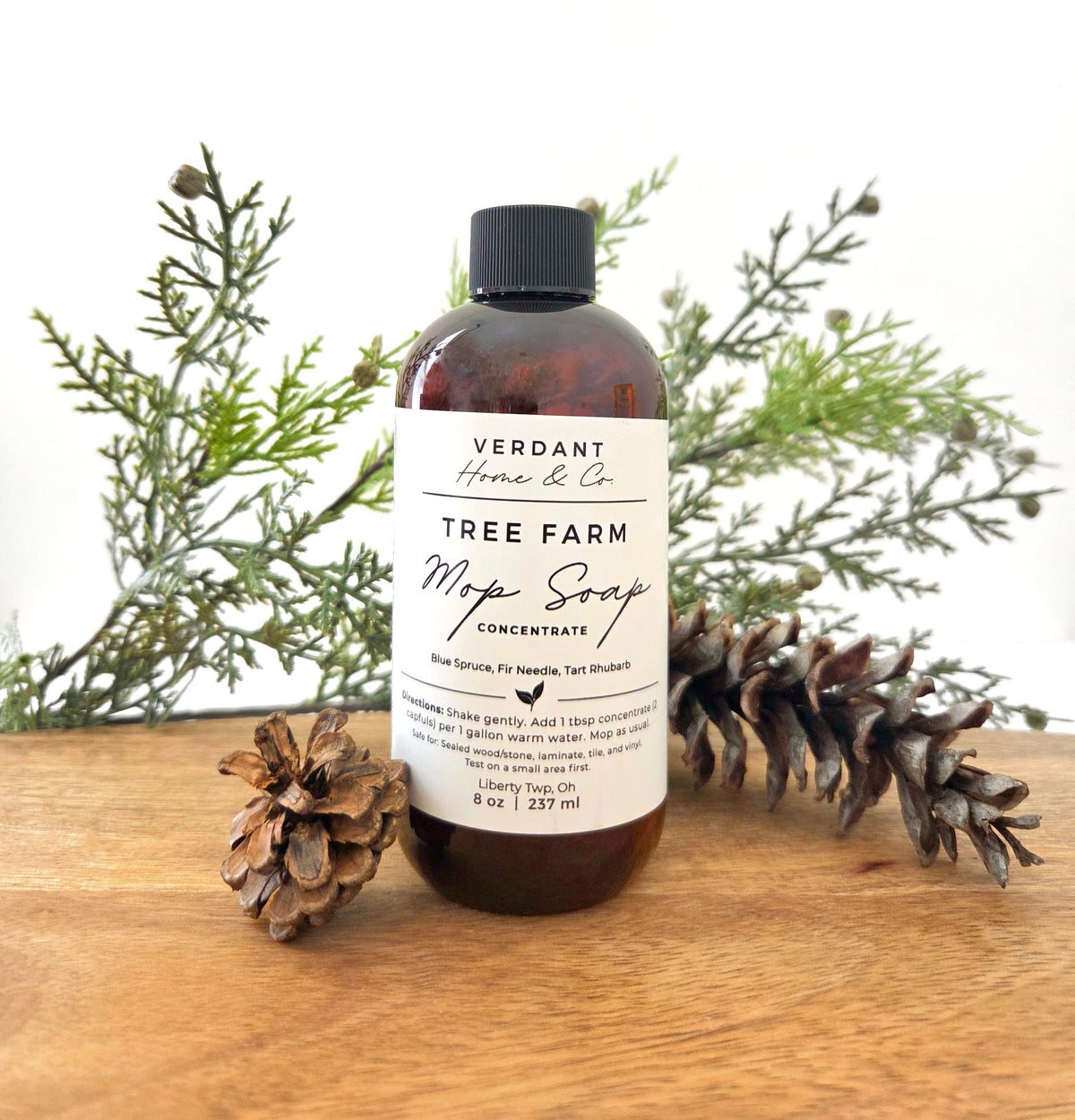 Bottle of Verdant Home & co mop Soap, in Tree Farm scent, on a wooden base with pinecones on either side.