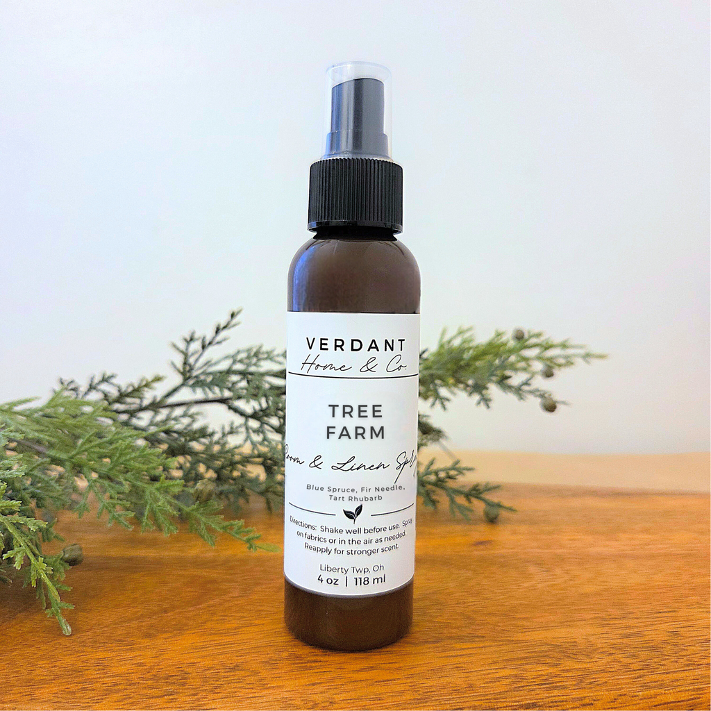 Bottle of Verdant Home & Co Tree Farm Room & Linen Spray on a wooden surface with greenery in the background.