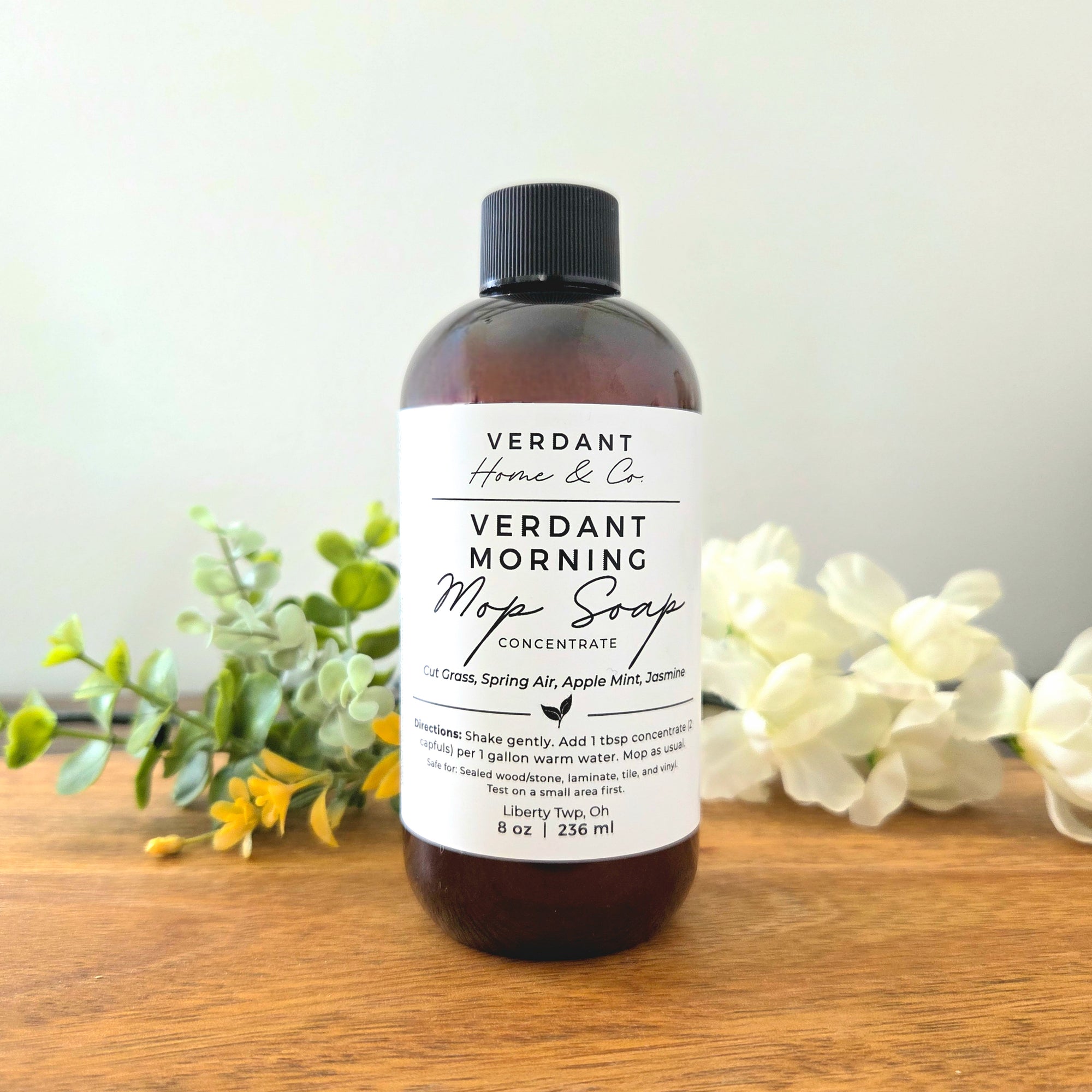 Bottle of Verdant Morning Mop Soap on a wooden surface with flowers and greenery.