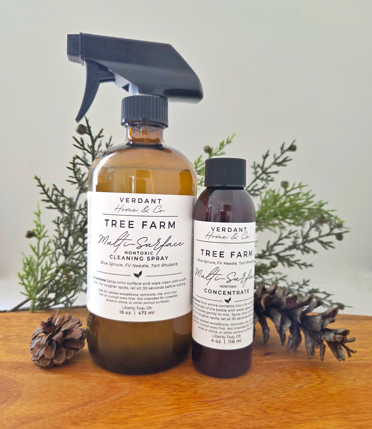 Two bottles of Verdant Home & Co Tree Farm Multi-Surface Cleaning Spray on a wooden surface with plants in the background.